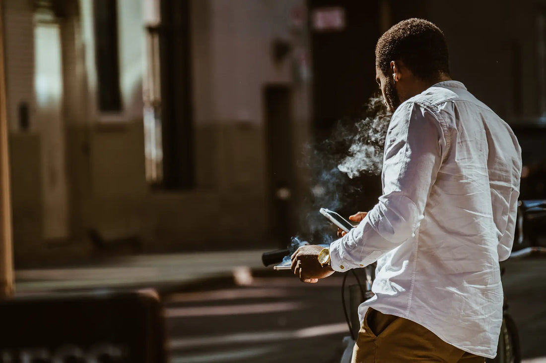 Mann steht mit dem Rücken zur Kamera, ein Smartphone in der rechten, eine Zigarette in der linken Hand.