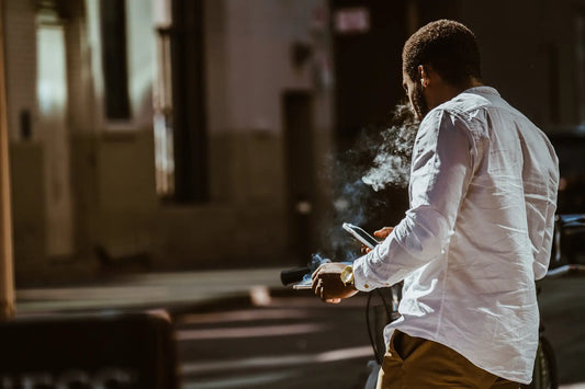 Mann steht mit dem Rücken zur Kamera, ein Smartphone in der rechten, eine Zigarette in der linken Hand.