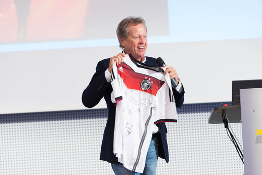 Klaus Eder, ehemaliger Physiotherapeut des DFB, hält auf dem ARTZT Symposium ein Fußballtrikot der Deutschen Nationalelf mit verschiedenen Unterschriften. | ARTZT neuro